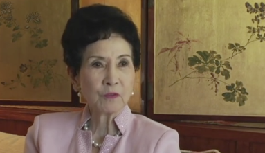Margaret Oda in a blush pink blazer and pearl necklace and earrings speaking to the camera